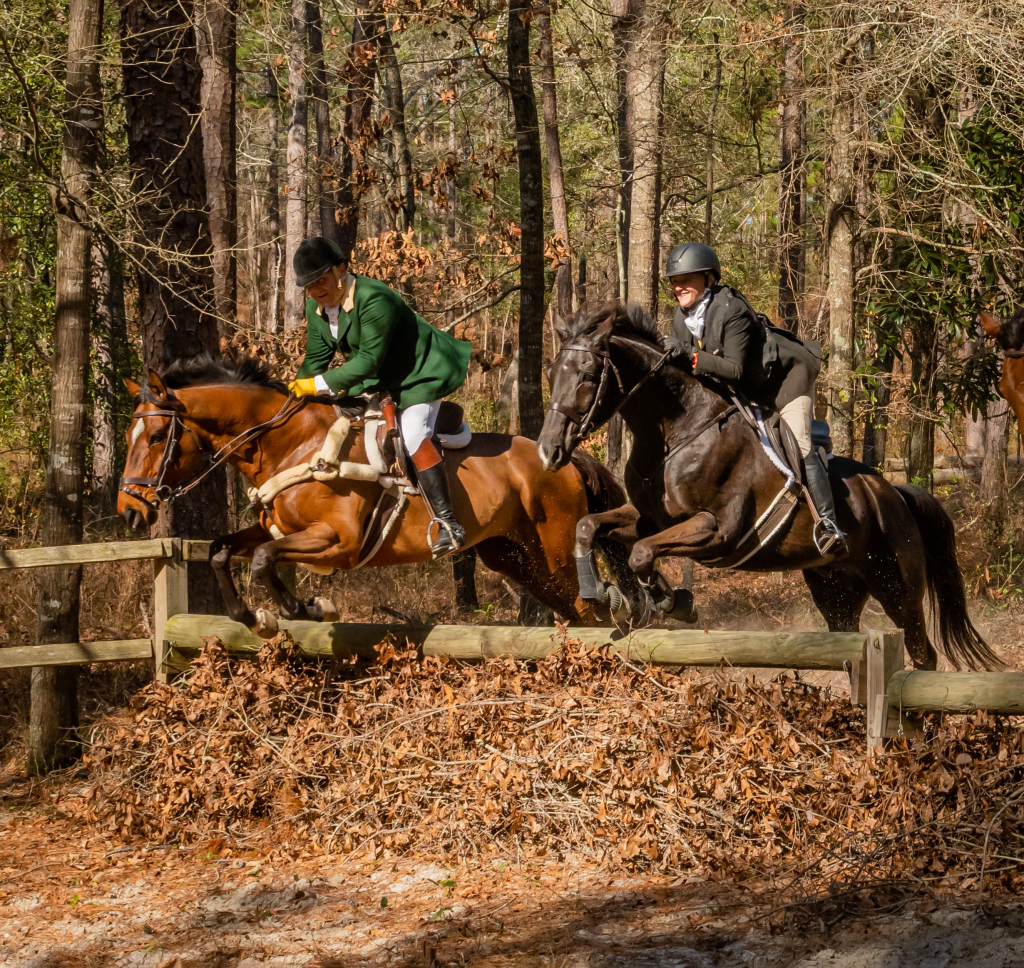 Two hunt riders jumping log fence side by side.