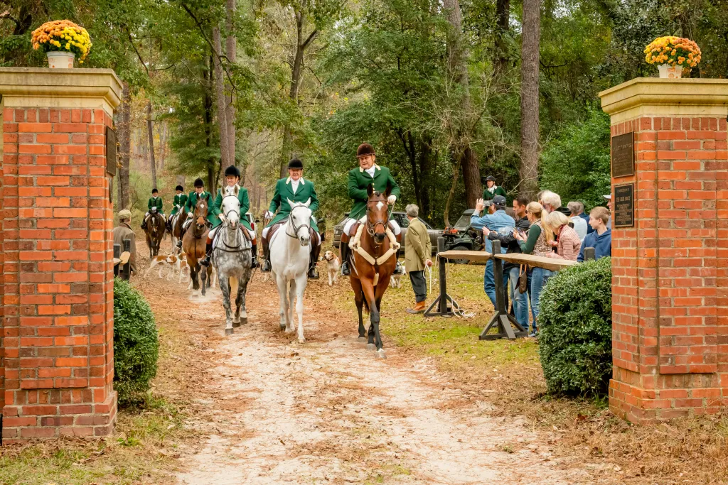 Hunt riders in green coats walking horses through brick pillars.