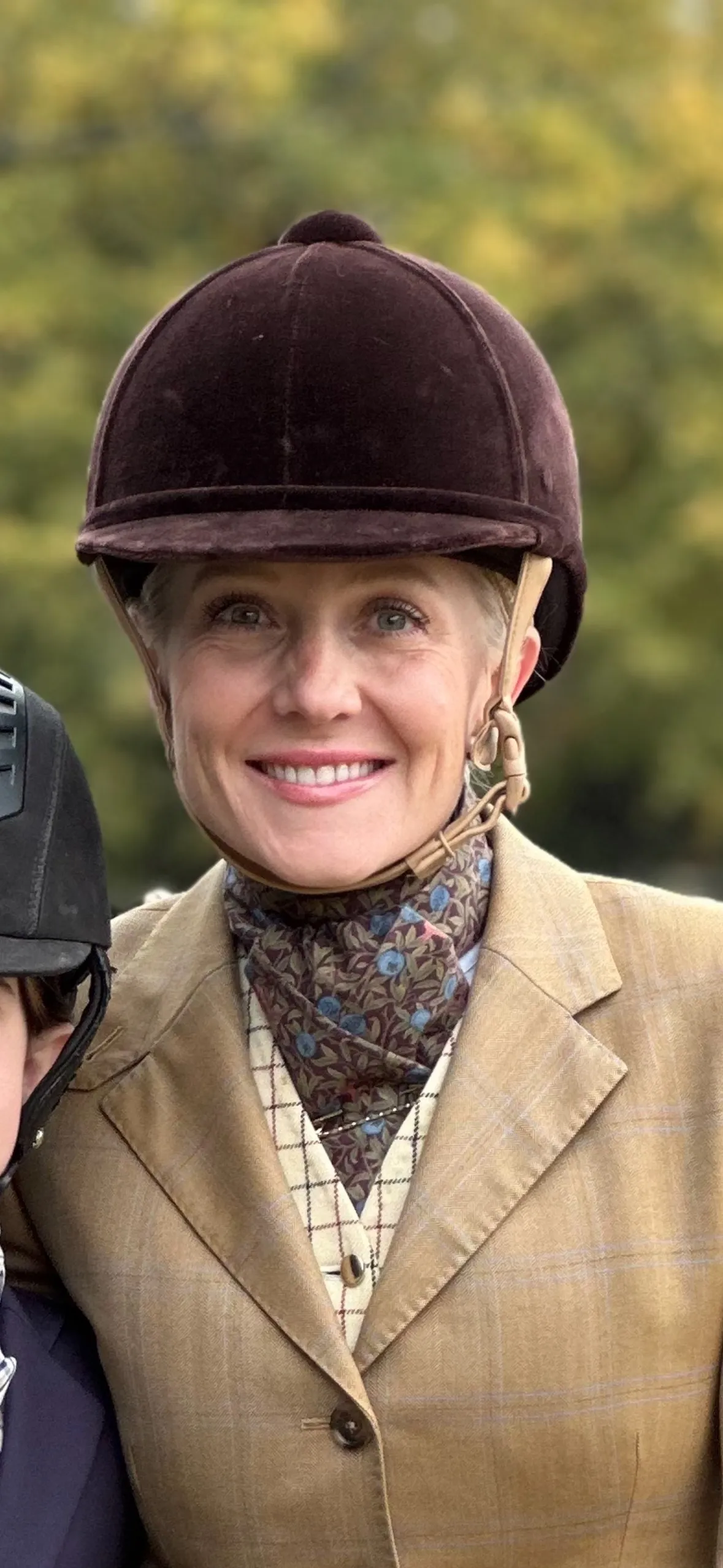 Woman in tan coat with vest and tie, brown suede helmet.